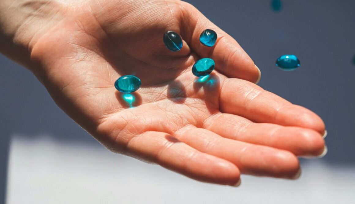 Hand holding several translucent blue liquid gel capsules falling into a palm. Supplement evaluation.