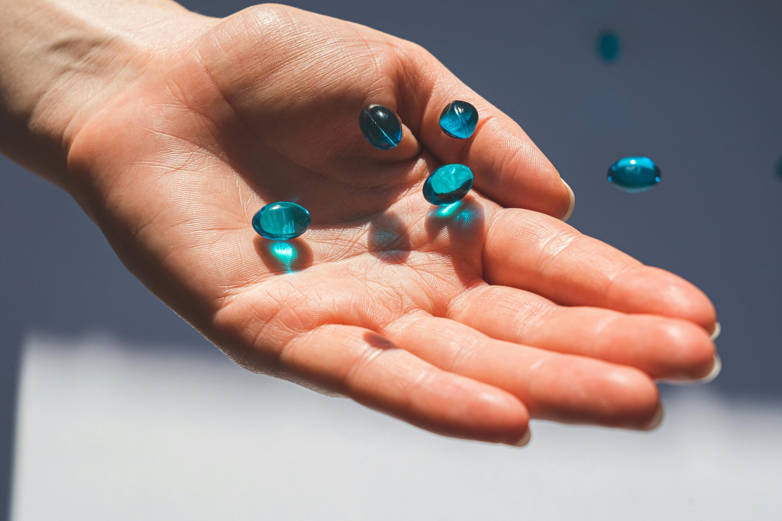 Hand holding several translucent blue liquid gel capsules falling into a palm. Supplement evaluation.