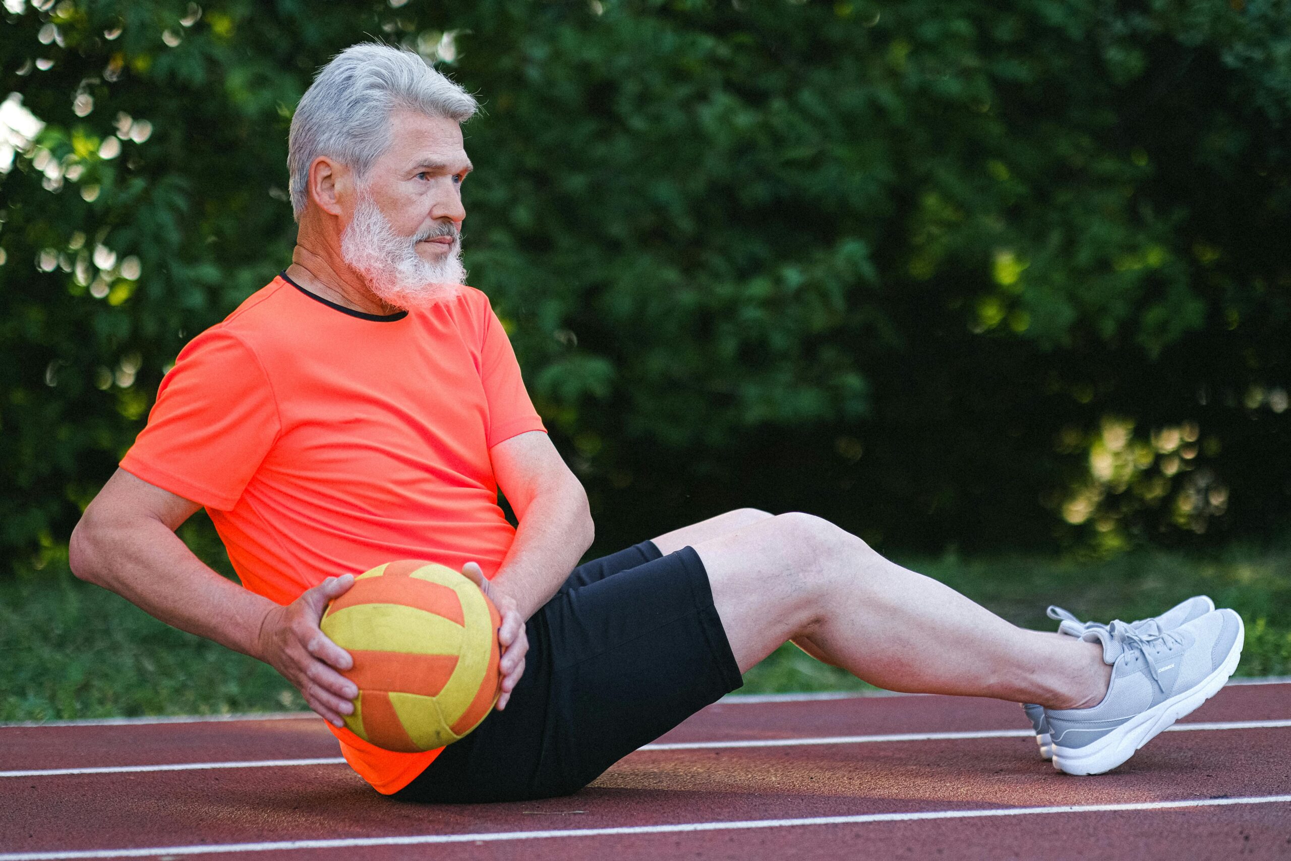 Senior man with a white beard performing a Russian twist core exercise with a medicine ball on an outdoor running track.