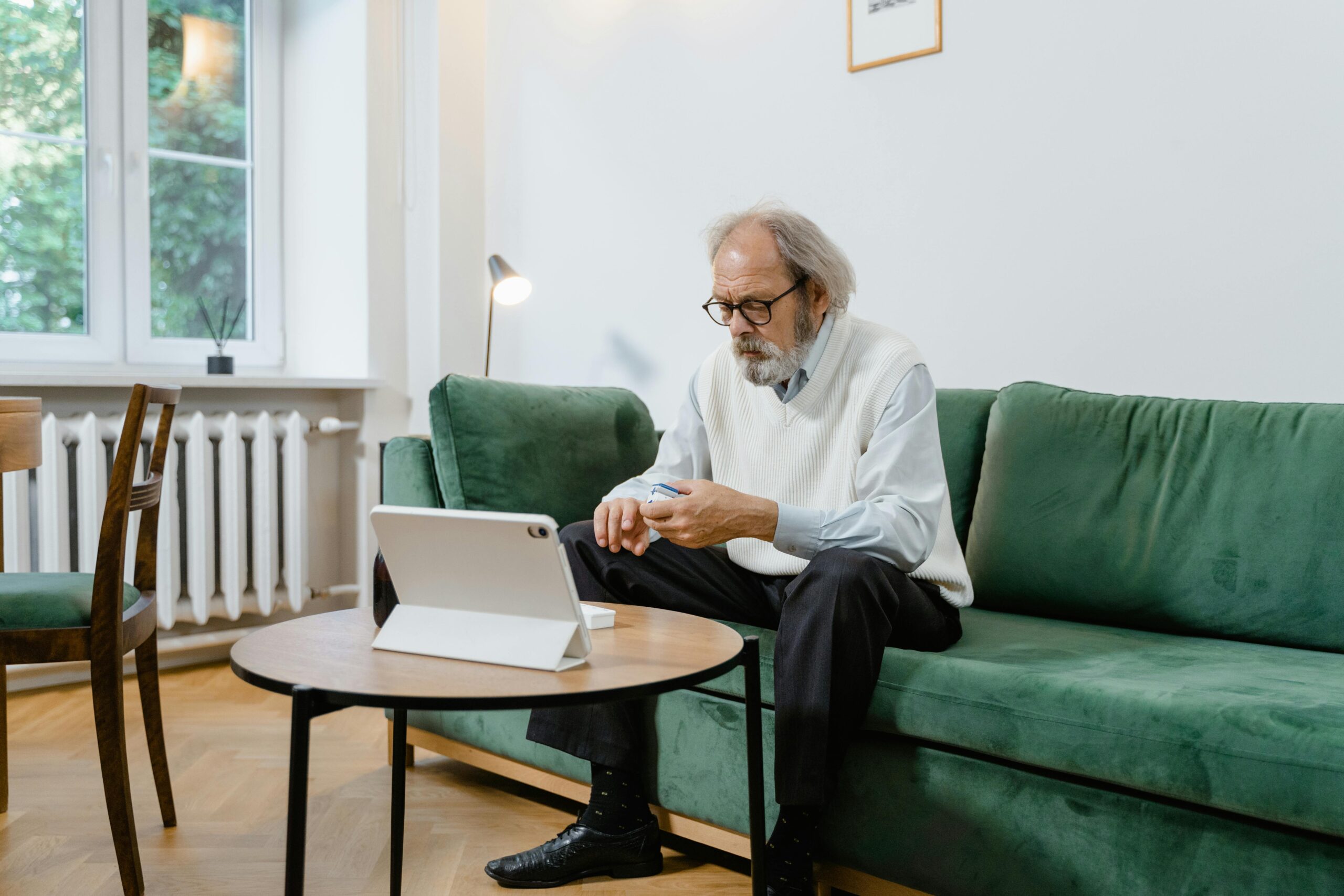 An older man over 50 sitting on a green sofa, holding a bottle of memory supplements and looking at a digital tablet to research brain health and cognitive support.