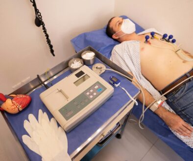A male patient lying on a medical exam table undergoing an electrocardiogram (ECG/EKG) test with chest electrodes and a pulse oximeter attached, next to a portable ECG monitor and a heart model.