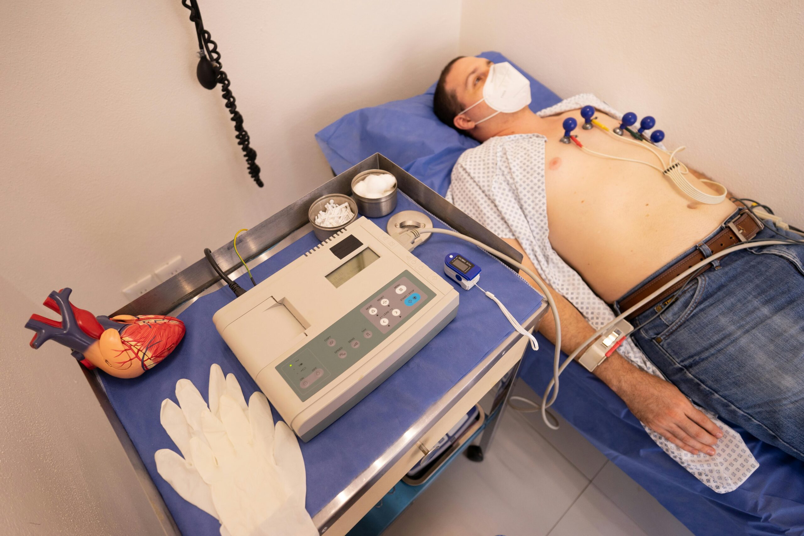 A male patient lying on a medical exam table undergoing an electrocardiogram (ECG/EKG) test with chest electrodes and a pulse oximeter attached, next to a portable ECG monitor and a heart model.