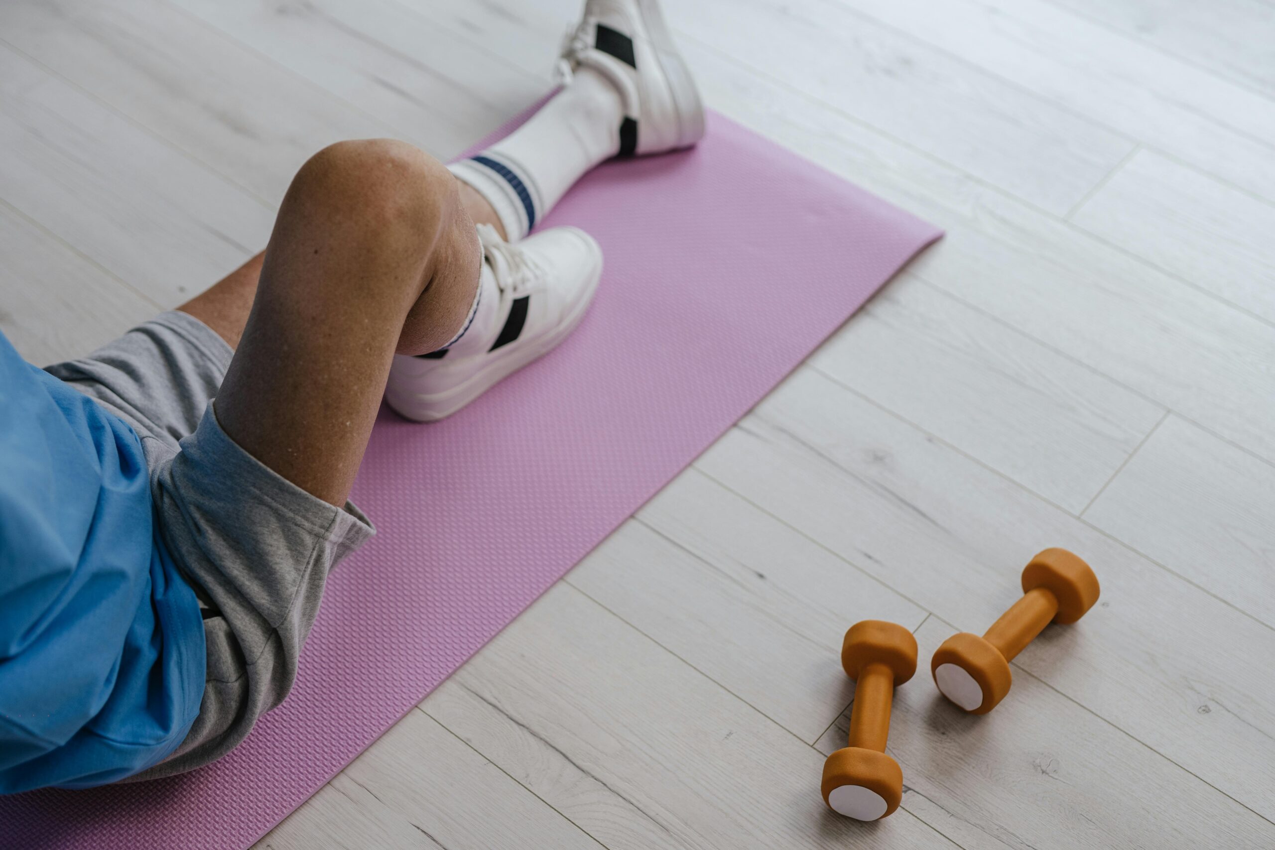 A senior man sitting on a pink yoga mat during a home workout, with two orange dumbbells on the floor.