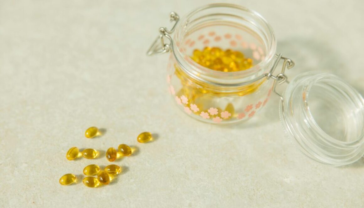 Yellow liquid softgel capsules spilled on a neutral stone countertop next to an open decorative glass jar.