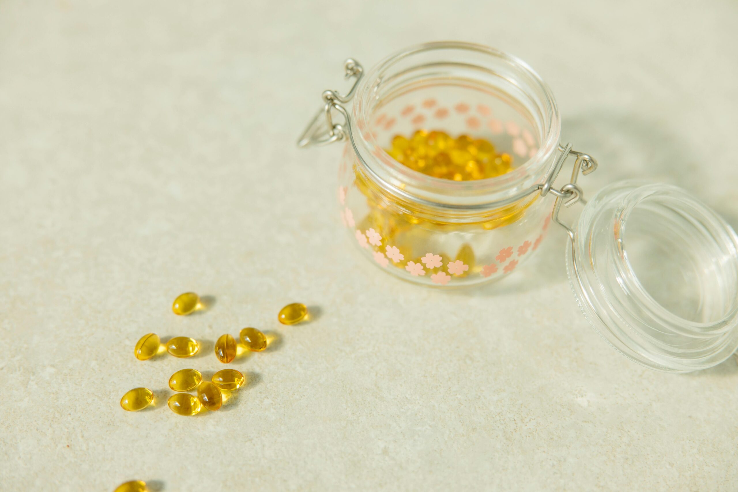 Yellow liquid softgel capsules spilled on a neutral stone countertop next to an open decorative glass jar.