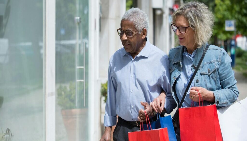 An older Black man and an older white woman walking together on a sunny sidewalk, carrying several colorful shopping bags while looking at a storefront window.