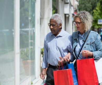 An older Black man and an older white woman walking together on a sunny sidewalk, carrying several colorful shopping bags while looking at a storefront window.