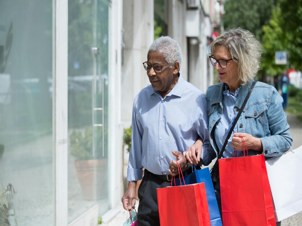 An older Black man and an older white woman walking together on a sunny sidewalk, carrying several colorful shopping bags while looking at a storefront window.