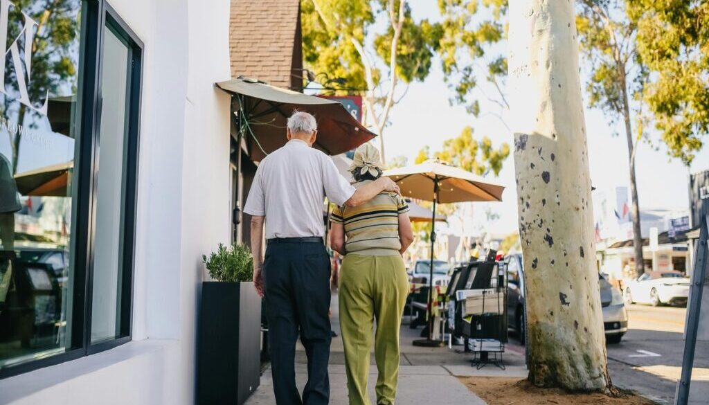 An elderly couple walks away from the camera down a sunny, tree-lined sidewalk. The man has his arm draped affectionately around the woman's shoulder as they stroll past white storefronts and outdoor cafe umbrellas.