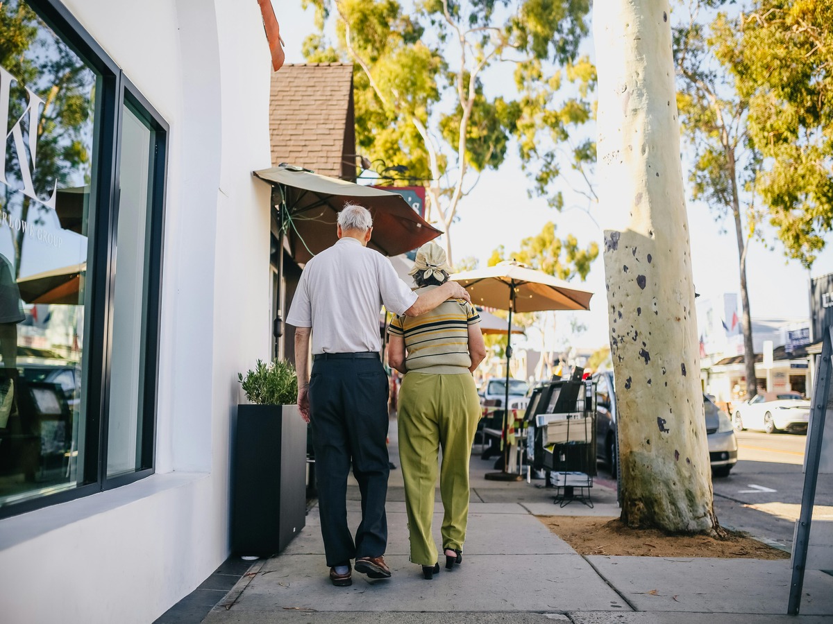 An elderly couple walks away from the camera down a sunny, tree-lined sidewalk. The man has his arm draped affectionately around the woman's shoulder as they stroll past white storefronts and outdoor cafe umbrellas.