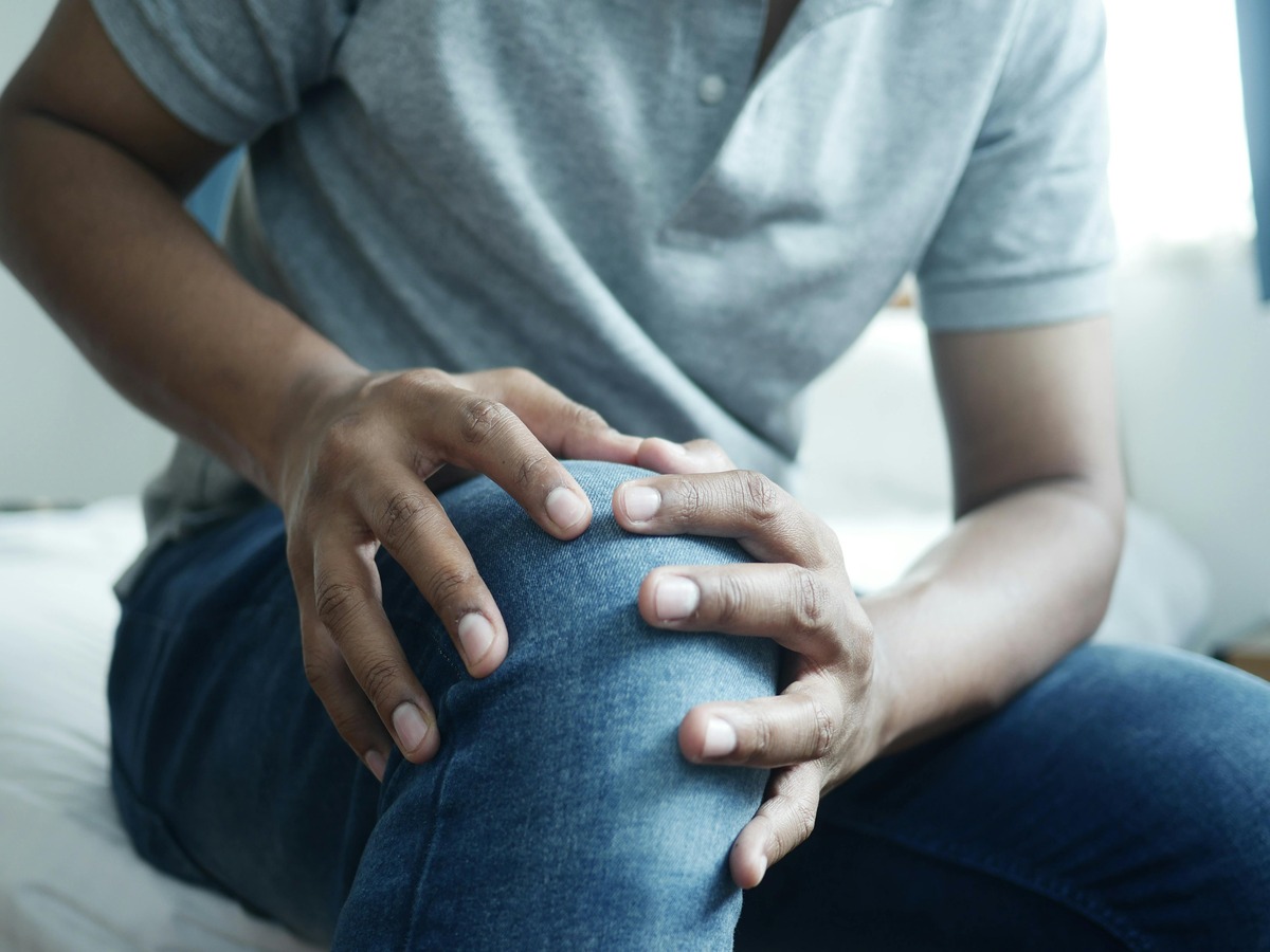 A person sitting on a bed, wearing jeans and a grey polo shirt, holding their knee with both hands in a gesture of discomfort and pain.