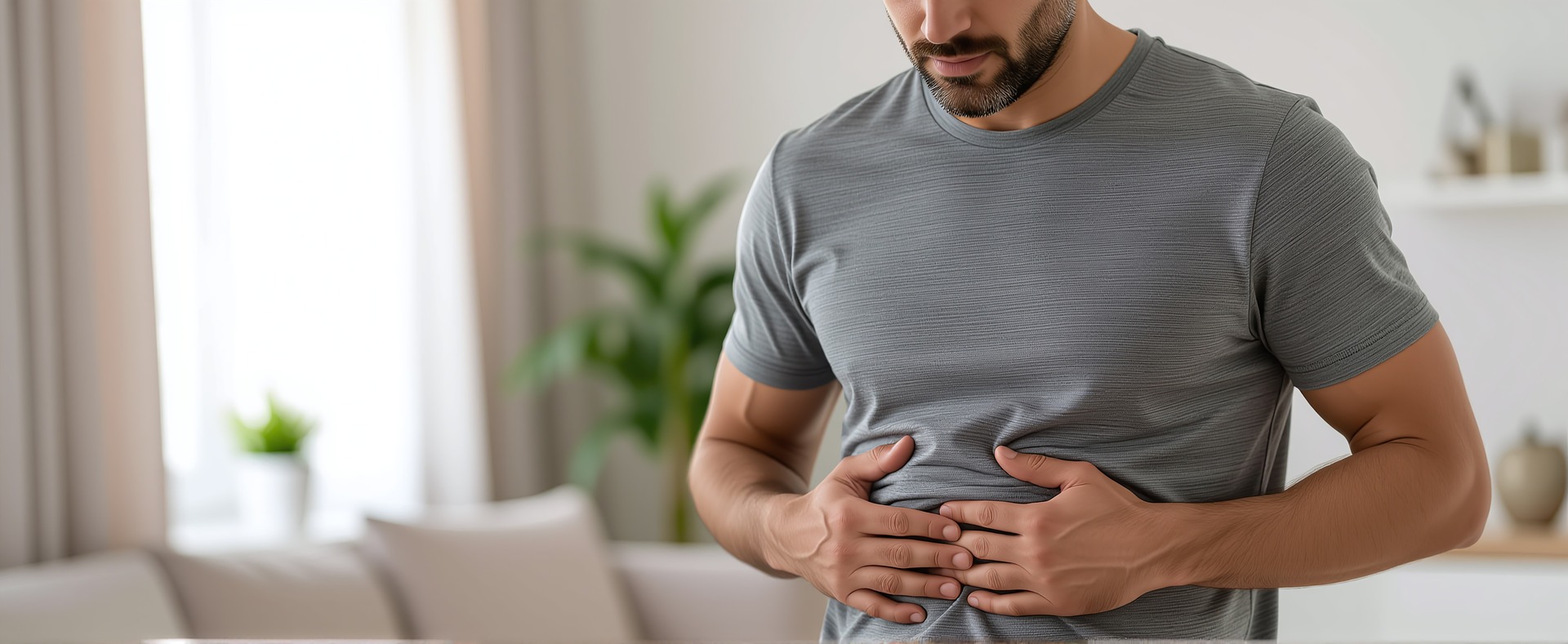 A close-up of a man in a gray t-shirt holding his stomach with both hands, suggesting abdominal discomfort or bloating. He is indoors with a softly blurred living room background.