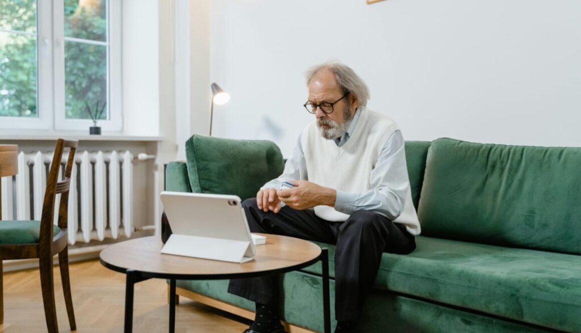 An older man with a gray beard and glasses sitting on a green couch, looking at a pulse oximeter on his finger while consulting a tablet on the coffee table.