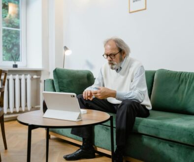 An older man with a gray beard and glasses sitting on a green couch, looking at a pulse oximeter on his finger while consulting a tablet on the coffee table.